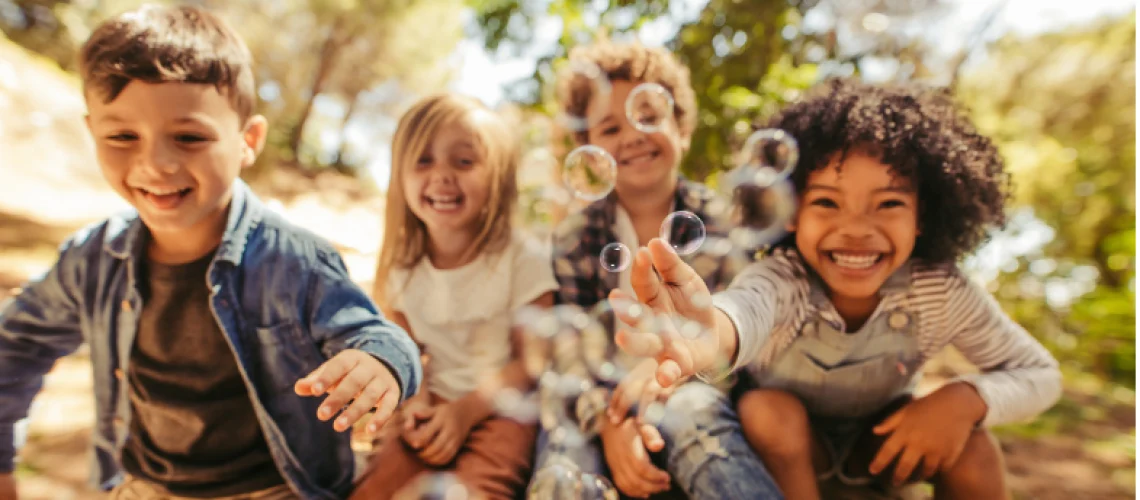 Four children smiling and reaching for bubbles outdoors.