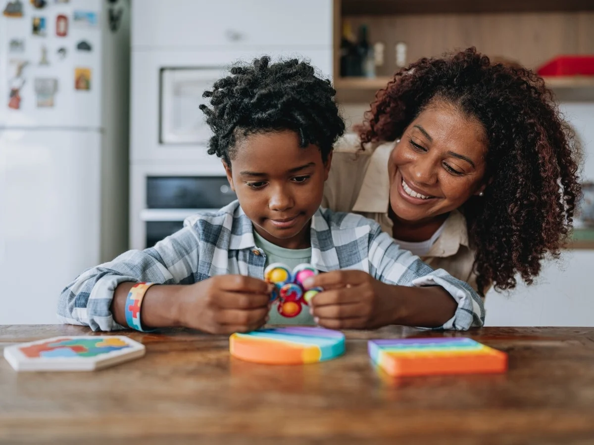 An adult smiling beside a child as they use a colourful sensory toy at a table.